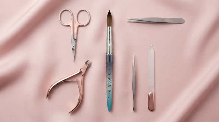 Collection of high-quality nail nippers and cuticle scissors with rose-gold accents, styled elegantly on a pastel pink silk surface, featuring a Kolinsky nail brush.